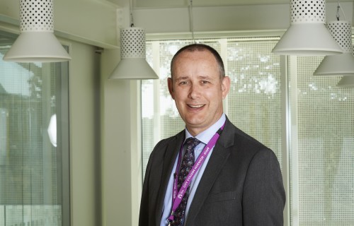 A teacher stands in a classroom smiling at the camera